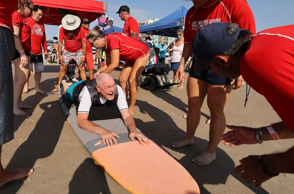 Volunteers help Navy veteran Rick Rettinger (bottom) onto a surfboard...