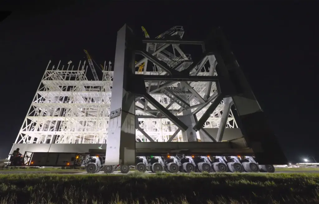 A steel truss is transported in front of SpaceX's under-construction...
