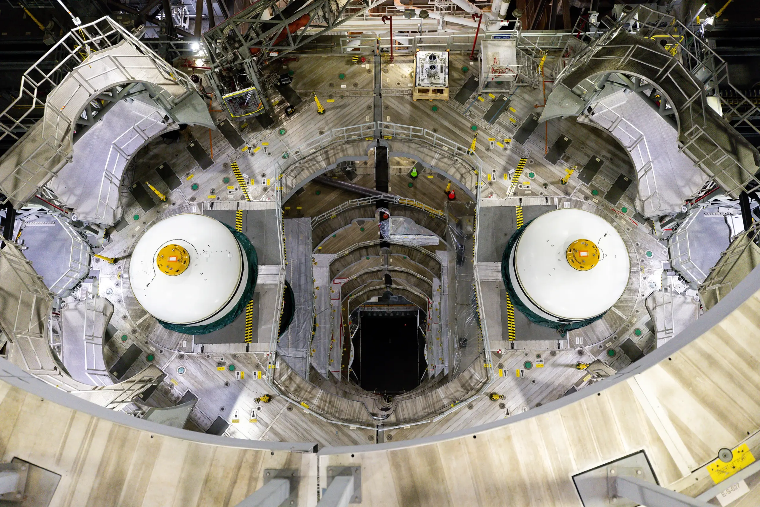 A top-down view inside a large rocket assembly bay, showing two cylindrical rocket sections with white caps positioned on either side of a central work area. Surrounding platforms, scaffolding, and safety railings form a circular pattern around the hardware, with tools, cables, and caution markings visible throughout the brightly lit workspace.